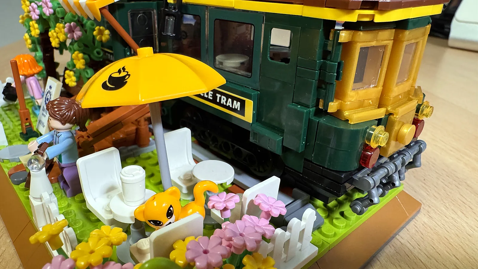 Detail des CraftBrickWorks Vintage Train Café mit Katze, gelbem Sonnenschirm und Gitarristin vor dem grünen Tram